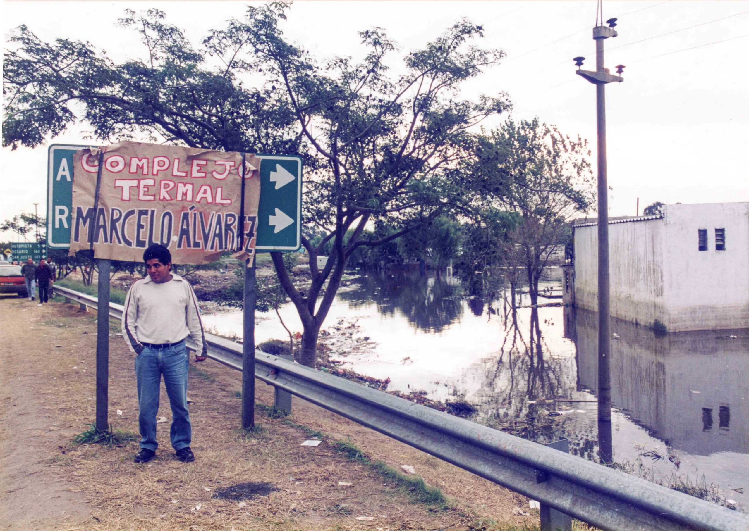 Persona frente a un cartel vial intervenido, donde puede leerse "Complejo Termal Marcelo Álvarez"